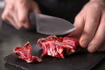 Man cutting raw meat on slate plate in kitchen, closeup