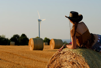 A young woman dressed as a cowgirl pauses on a  hay bale and enjoys the sunset and the new way of...