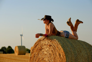 A young woman dressed as a cowgirl pauses on a  hay bale and enjoys the sunset and the new way of...