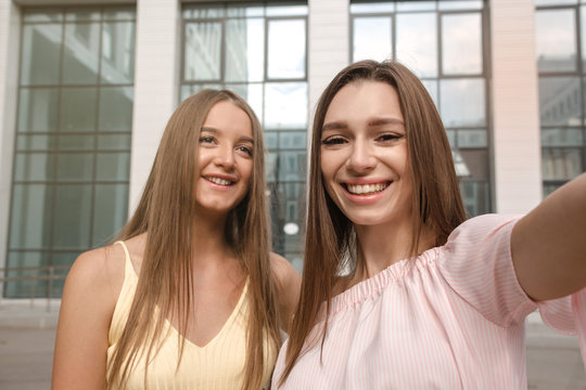 young girls take pictures of themselves on the camera smartphone in short shorts and blouses, smiling, against the background of a multi-storey building