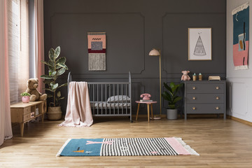 Real photo of a baby crib in a grey child's room interior, next to a cupboards, lamp and plants - obrazy, fototapety, plakaty
