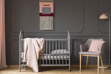 Real photo of a baby crib with a blanket standing next to an armchair with a cushion and a lamp in child's room interior - obrazy, fototapety, plakaty