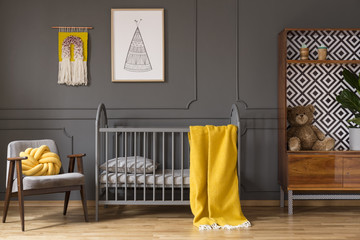 Real photo of a baby cot with yellow cot standing between an armchair and a cupboard with teddy...