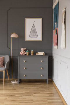 Real photo of a grey cupboard with ornaments standing next to a lamp, armchair and wall with a poster in spacious baby room interior