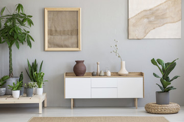 Posters on grey wall above white cupboard in living room interior with plants and pouf. Real photo