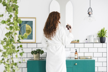 Woman in front of mirror in white bathroom interior with poster, plants and green cabinet. Real photo