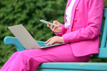 Young woman with laptop and mobile phone resting on bench outdoors