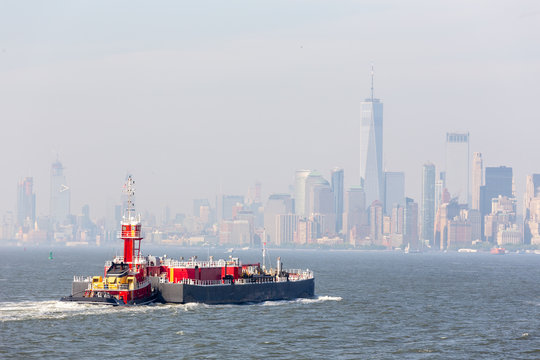 Freight tug pushing cargo ship to the port in New York City and Lower Manhattan skyscarpers skyline in background. New York City, USA.