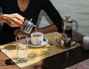 A woman's hand is pouring fresh milk from a pot into a coffee cup, with a French press and metal set, in a restaurant.