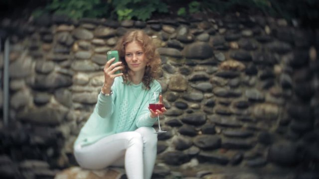 A woman is using a smartphone against a back wall. Drinks and drinks wine from a glass