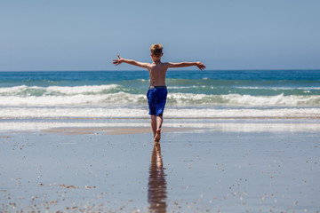 A young boy is walking away from the beach into the ocean with his arms up