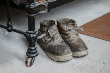 old-fashioned leather shoes stand on a concrete floor next to an old metal bed.