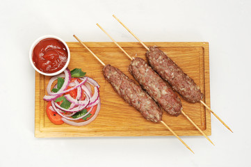 Lulya kebab on wooden skewers. Next to a kebab salad of ripe tomato and slices of red onion. In a cup, sauce is ketchup. White background. Close-up. View from above. 