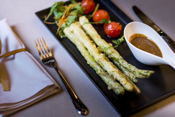 Deep fried asparagus with a dipping sauce and tomatoes on black plate. Selective focus