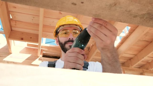Portrait of a male builder in overalls and a yellow helmet on the roof of a frame house. Screws with a screwdriver.