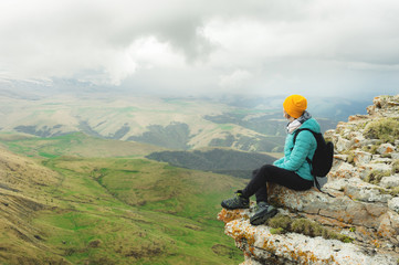 Young woman with a backpack pensively sitting on the edge of a rock and looking at the sky with clouds