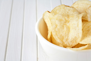 Delicious golden potato chips in a white bowl on a white wooden background.