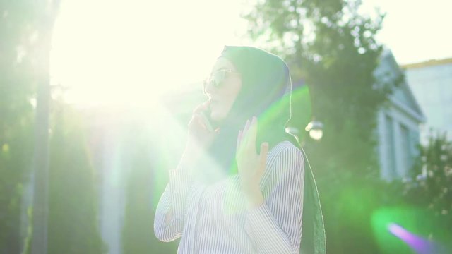Young stylish attractive muslim woman in hijab and glasses talking on the phone,sunflare.Slow mo