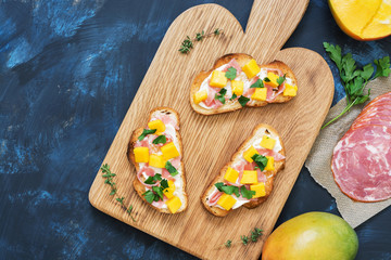 Sandwiches with cream cheese, ham and mango on a wooden board, blue background. The view from above, flat lay.