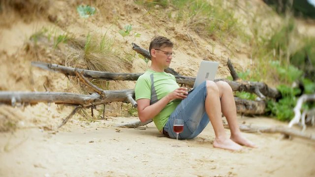 busy mature man working on a laptop computer with business papers picking up a phone to work a call sitting on the beach by the sea on a sunny day and drinking wine from a glass