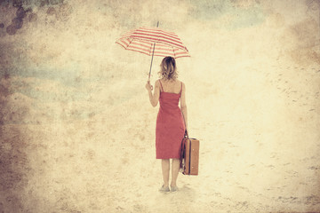 Young woman in red dress with umbrella and suitcase on the beach. Travel concept image on sand. Photo in old color image style.