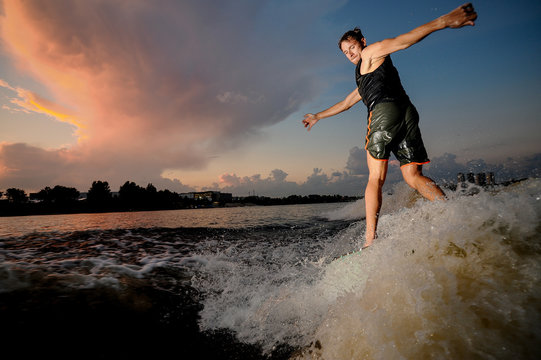 Active athletic man riding on wakesurf down the river during sunset