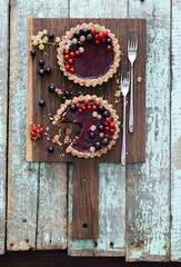 Small vegan tarts made of nuts and berry jam decorated with black, red and white currants on oak cutting board