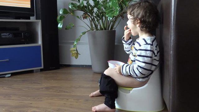 Cute little boy eating snack and watching tv while sitting on the potty
