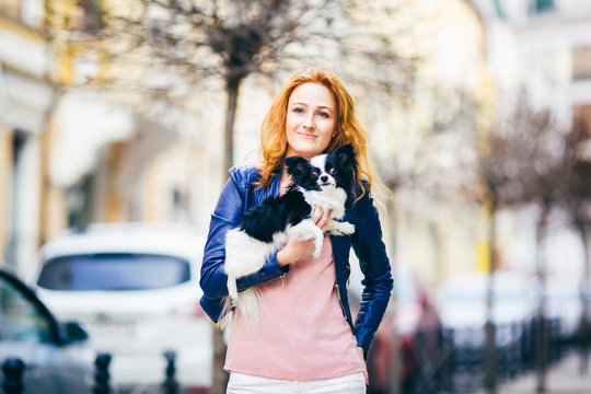 Subject man and dog. young red-haired Caucasian woman with freckles on face holds black and white shaggy chihuahua breed dog. The girl dressed in blue leather jacket, stands on busy street in spring