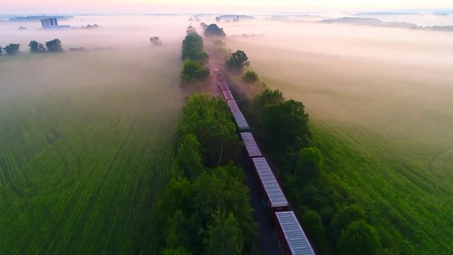 Freight train rolls across foggy rural landscape at sunrise with ethereal beauty, aerial view.