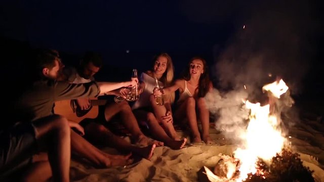 Beach party at sunset with bonfire. Friends sitting around the bonfire, drinking beer and singing to the guitar. Young men and women hold glass bottles with beverage singalong and cheering.