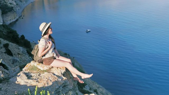 Modest young female traveler with backpack sitting on cliff and playing her bare feet at sunset