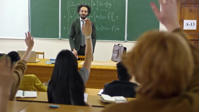 Tracking shot of multi ethnic group of university students seen from their back raising hands to answer teacher’s questions during lecture on mathematics