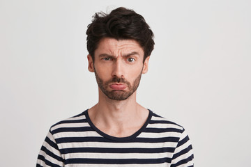 Closeup of sad offended young man with bristle wears striped t shirt feels upset and looks directly in camera isolated over white background