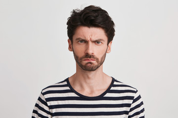 Portrait of angry displeased young man with bristle wears striped t shirt looks serious and irritated isolated over white background