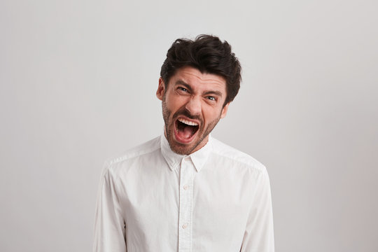 Indoor shot of young unshaven businessman screams and feel mad and angry, because the project isn't done. Negative facial expression. Isolated over white wall