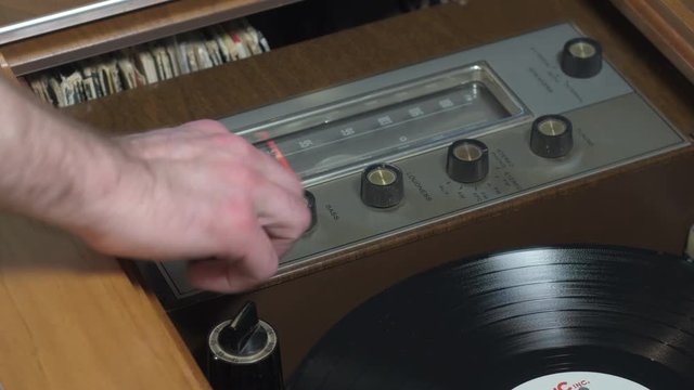 Hand turning on a retro wooden cabinet stereo and record player. A vinyl record automatically drops onto the turntable and starts spinning.