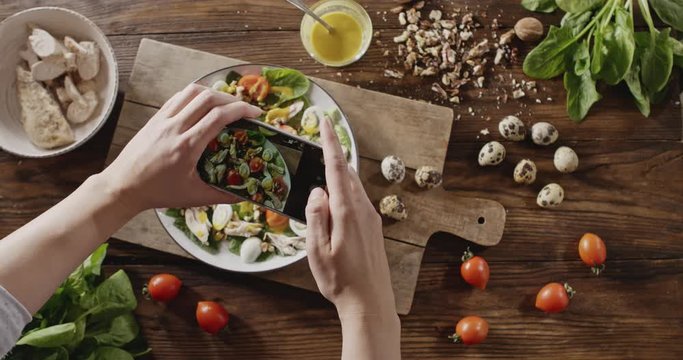 A woman's hands make photos mobile phone of salad from natural ingredients. Top view of background with quail eggs, chicken fillet, greens, tomatoes, walnut for cooking salad. Slow motion video in 4K