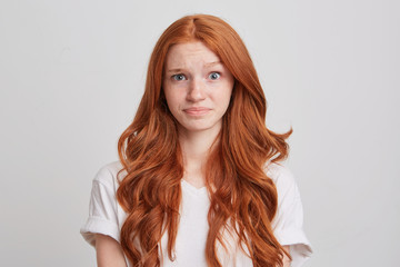 Portrait of confused astonished young woman with long wavy red hair and freckles wears t shirt feels embarrassed and looks directly in camera isolated over white background - obrazy, fototapety, plakaty