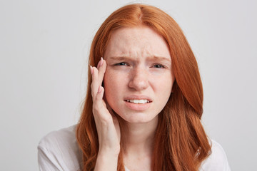 Closeup of upsed depressed young woman with long wavy red hair and freckles touching her temple and...