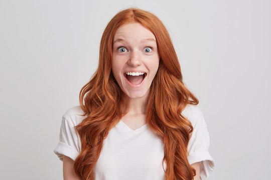 Portrait of happy excited redhead young woman with long hair and freckles wears t shirt looks surprised and impressed isolated over white background