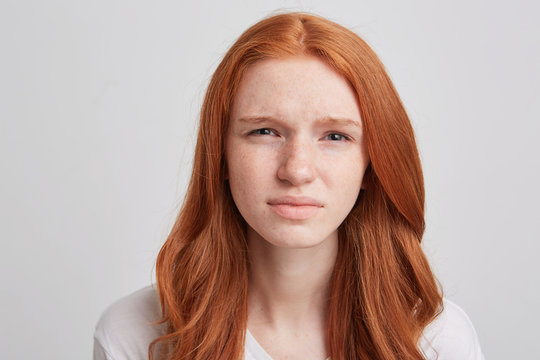 Portrait of sad unhappy young woman with long wavy red hair and freckles feels upset and looks directly in camera isolated over white background