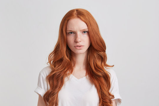 Closeup of thoughtful beautiful redhead young woman with long wavy hair wears t shirt looks directly in camera and thinking isolated over white background