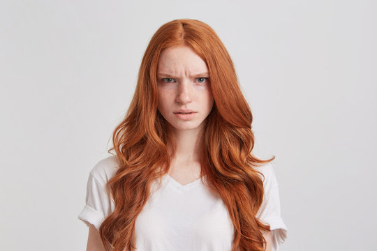 Portrait of angry frowning young woman with long wavy red hair wears t shirt looks mad and irritated isolated over white background