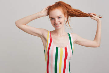 Portrait of happy beautiful young woman with red hair and freckles in striped top holding her ponytail and posing isolated over white background Looks satisfied - obrazy, fototapety, plakaty