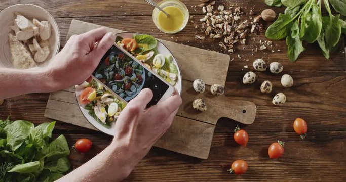 A woman's hands make photos mobile phone of salad from natural ingredients. Top view of background with quail eggs, chicken fillet, greens, tomatoes, walnut for cooking salad. Slow motion video in 4K