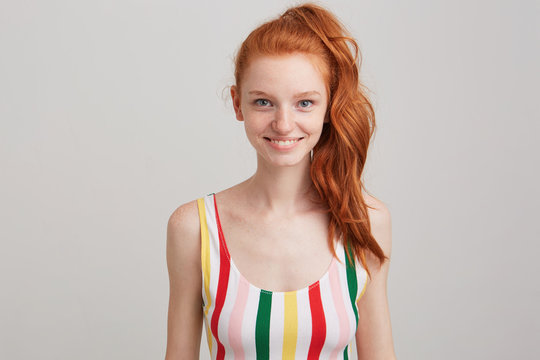 Closeup of smiling attractive redhead young woman with freckles and ponytail wears striped top feels confident and looks directly in camera isolated over white wall