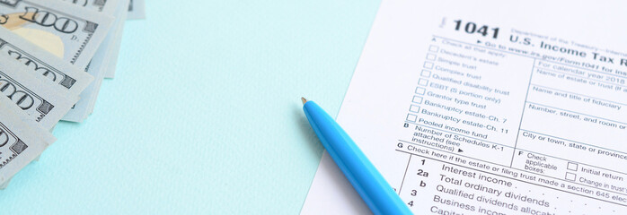 1041 tax form lies near hundred dollar bills and blue pen on a light blue background. US Income tax...