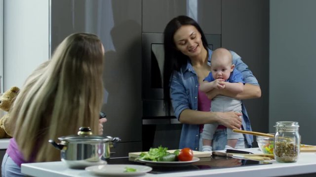 Medium shot of young woman with long hair holding mobile phone and taking picture of her female partner and baby boy in kitchen