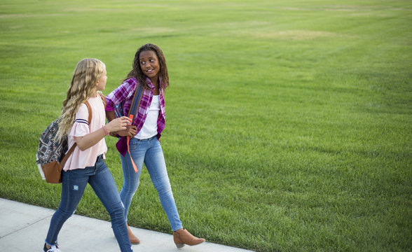 Two Diverse School Kids Walking And Talking Together On The Way To School. Back To School Photo Of Diverse Girls Wearing Backpacks In The School Yard